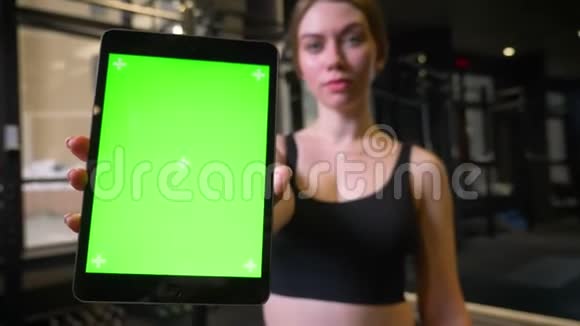 扎着马尾辫的女运动员的特写镜头显示在健身房里带有彩色绿色屏幕的平板电脑是积极的视频的预览图