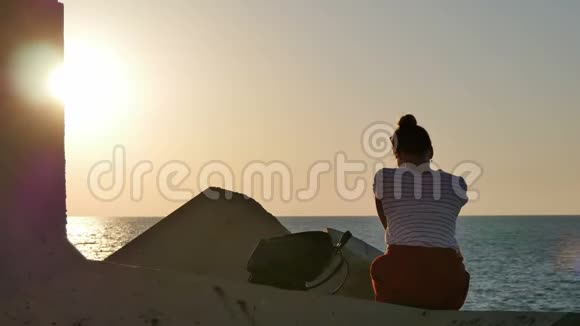 梦想着日落时分坐在海边的岩石上的年轻女子视频的预览图