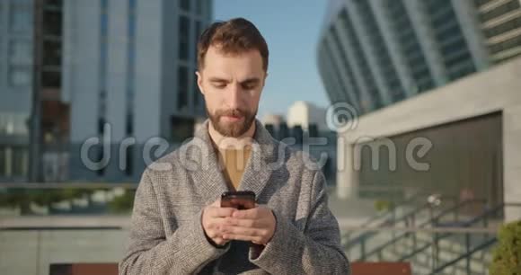 留胡子的时髦千禧一代穿着外套的男人在手机上聊天看着镜头的肖像手持射击视频的预览图
