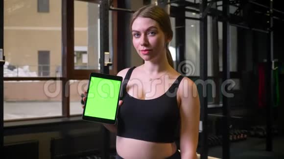 扎着马尾辫的女运动员将带有彩色绿色屏幕的平板电脑展示成相机在健身房里保持冷静和微笑视频的预览图