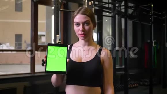 扎着马尾辫的女运动员展示了带有彩色绿色屏幕的平板电脑在健身房里保持冷静和积极视频的预览图