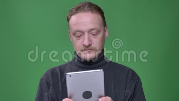 身着灰色套头衫的金发养老金领取者专注地看着平板电脑并对绿色背景做出情感反应视频的预览图