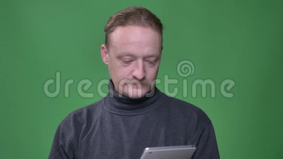 穿灰色套头衫的金发养老金领取者专心致志地工作在平板电脑上并在绿色背景上展示了它的蓝色屏幕视频的预览图