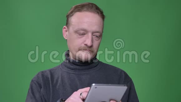 穿灰色套头衫的金发养老金领取者专心地工作在平板电脑上并在绿色背景上观看相机视频的预览图