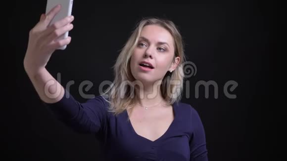 穿着蓝色短衫的中年金发女商人在黑色智能手机上在相机上笑着自拍视频的预览图