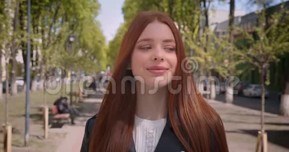 美丽的姜姑娘穿着皮夹克在绿城小巷的背景下漂亮地看着镜头视频的预览图