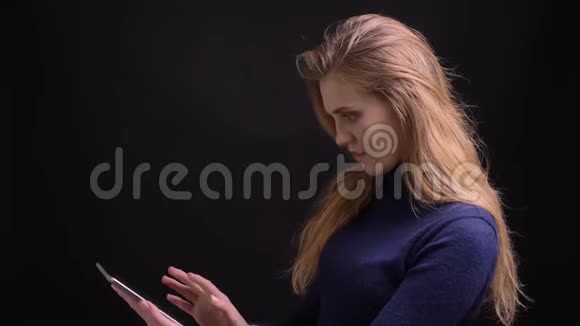 年轻的金发模特打开平板电脑上的应用程序在黑色背景下快乐地展示它的绿色屏幕视频的预览图