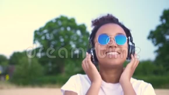 非洲裔美国少女穿着白色T恤和蓝色太阳镜听耳机音乐的年轻女性视频的预览图