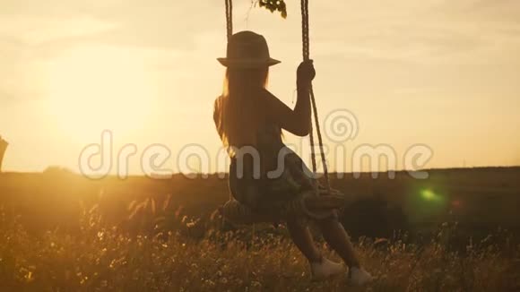 快乐的小女孩在金色的夏日夕阳下摇摆一个年轻的少女在孤树上摇摆的剪影视频的预览图