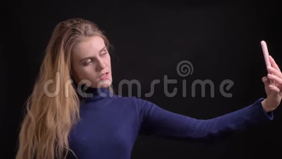 金发模特在黑色背景下在智能手机上调情和美丽的自拍照片视频的预览图