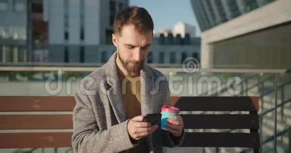 城市里坐在长椅上浏览和在手机上聊天的留胡子的时髦千禧一代男人的肖像手持视频的预览图