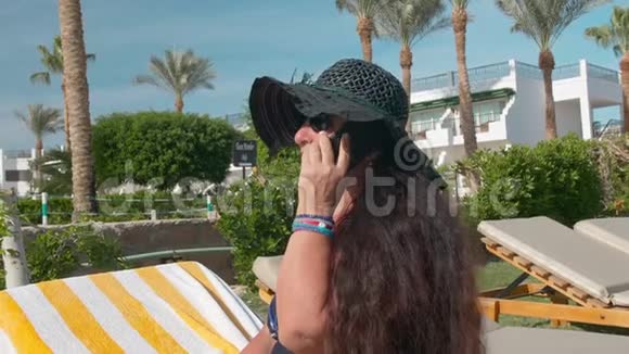 戴着黑帽子戴着智能手机坐在躺椅上晒太阳的白人老年女性视频的预览图