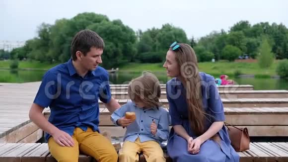 幸福家庭概念母亲父亲和小男孩坐在河边吃羊角面包视频的预览图