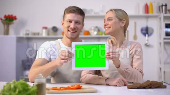 年轻的微笑夫妇展示绿色屏幕平板电脑烹饪课程模板视频的预览图