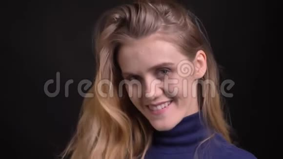 年轻的金发模特穿着蓝色毛衣观看镜头在黑色背景上调情和神秘的微笑视频的预览图