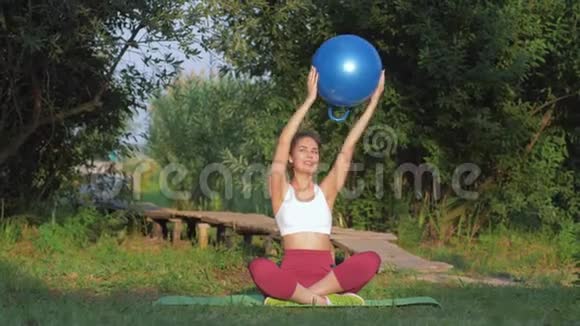 户外健身运动年轻女子在草地上的垫子上坐着一个大健身球锻炼手臂肌肉视频的预览图