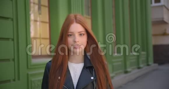 穿着皮夹克的红头女孩走在镜头前在绿色房子的背景上微笑着走进它视频的预览图