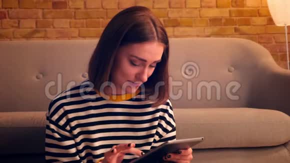 在一间舒适的公寓里年轻漂亮的女孩在平板电脑上打字开心地坐在地板上微笑特写照片视频的预览图