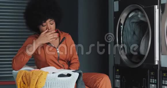 疲惫的年轻非洲裔美国妇女在洗衣店洗衣服时看书她打哈欠等待视频的预览图