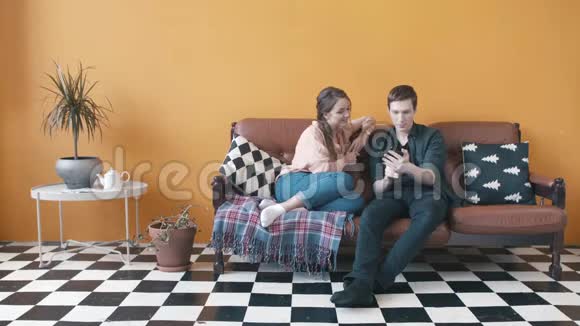 一对幸福的年轻夫妇坐在家里的沙发上的特写镜头男人在手机屏幕上显示一些东西股票视频的预览图