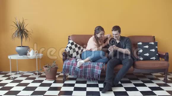 一对幸福的年轻夫妇坐在家里的沙发上的特写镜头男人在手机屏幕上显示一些东西股票视频的预览图