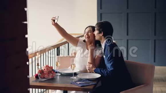 快乐的人男人和女人在餐馆里用智能手机摄像头自拍亲吻和大笑然后看着视频的预览图