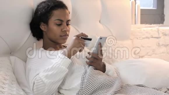 年轻的非洲女性在床上放松通过智能手机在线购物视频的预览图