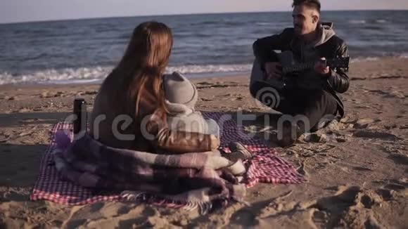 一家人坐在沙滩上的格子毯子上爸爸弹吉他妈妈带着孩子听他说话视频的预览图