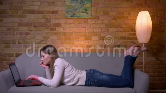 年轻有魅力的白种人深色头发的女性在笔记本电脑上上网躺在沙发上的特写照片视频的预览图