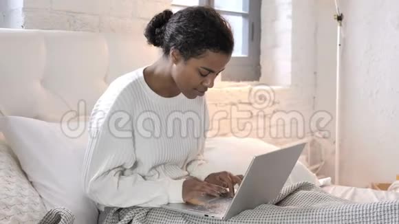 年轻的非洲妇女坐在床上庆祝笔记本电脑的成功视频的预览图