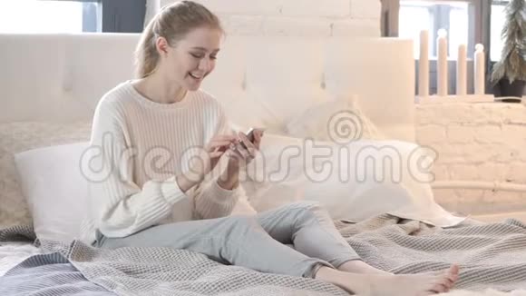 兴奋的女人坐在床上庆祝智能手机的成功视频的预览图