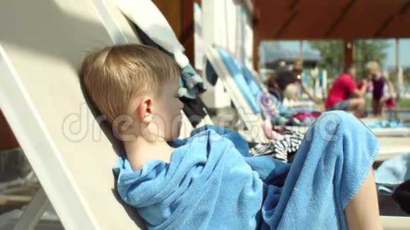 一个悲伤的小男孩在游泳池旁的休息室上的肖像他被裹在毛巾里视频的预览图