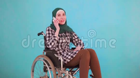 一位坐轮椅的年轻穆斯林妇女在蓝色背景下打电话视频的预览图