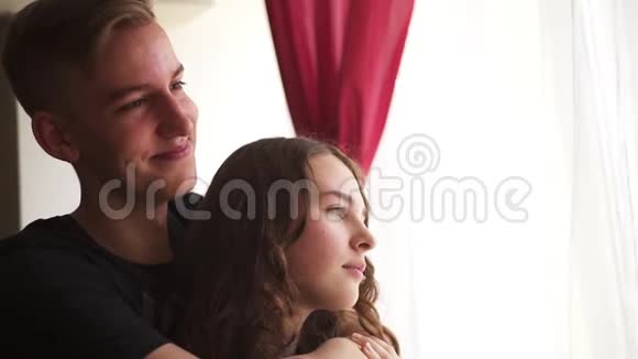 可爱的年轻人拥抱他的女朋友站在白天的窗户旁边视频的预览图