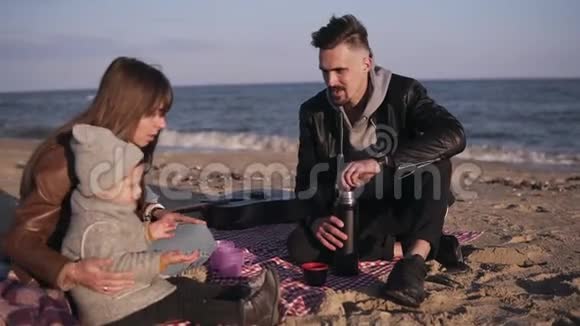 年轻的家庭穿着休闲装坐在水边的格子呢上野餐时喝热饮料寒冷的天气周末视频的预览图