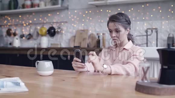 漂亮的年轻女人棕色头发穿着粉红色衬衫坐在厨房的木桌上端着茶或咖啡还有视频的预览图