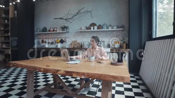 美丽的年轻女子坐在厨房的木桌旁喝茶或咖啡在她的银色笔记本电脑上打字视频的预览图
