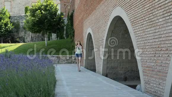 戴着一顶背包的年轻女人沿着古老的墙壁走着摸着她的手旅行和建筑视频的预览图