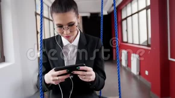 商业概念一个戴眼镜的女人坐在商务中心的秋千上听音乐看着视频的预览图
