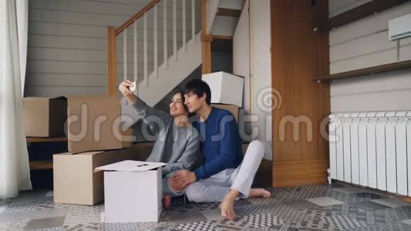 年轻人和女人正在用智能手机自拍用手指坐在新公寓的地板上做心形视频的预览图