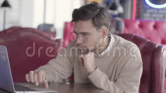 英俊的忧郁的年轻人带着眼镜看着笔记本电脑坐在咖啡馆或餐馆的桌子旁视频的预览图