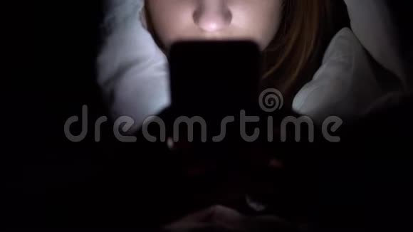 一个年轻的女人躺在毯子下手里拿着电话黑暗中的女人用垫子现代电子平板电脑视频的预览图