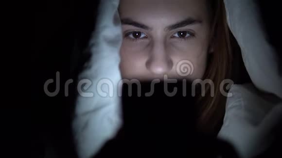 一个年轻的女人躺在毯子下手里拿着电话黑暗中的女人用垫子现代电子平板电脑视频的预览图