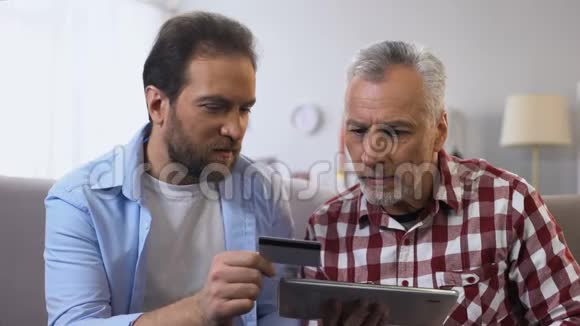 年轻的儿子和退休的父亲在标签上插入信用卡号码支付应用程序视频的预览图