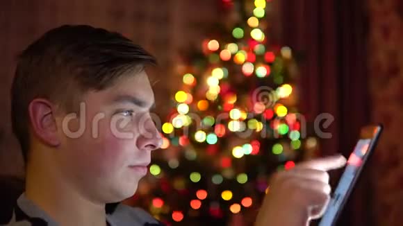 一个年轻人坐在圣诞树的背景上手里拿着一块平板电脑整个房间都是装饰的视频的预览图