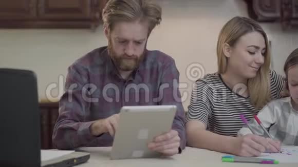 有胡子的人拿着平板电脑和笔记本电脑在桌子上工作那个女人坐在旁边小女孩帮着女儿视频的预览图