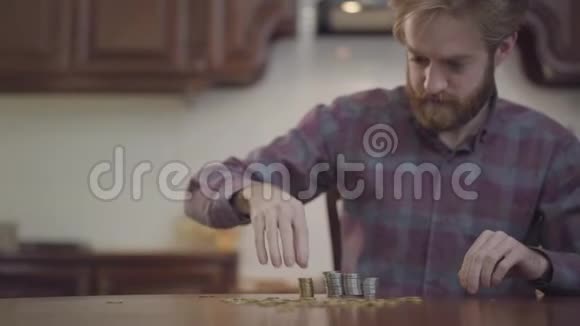 坐在厨房里数钱的桌子旁的留胡子男人的画像穿着格子衬衫的胡子堆硬币视频的预览图