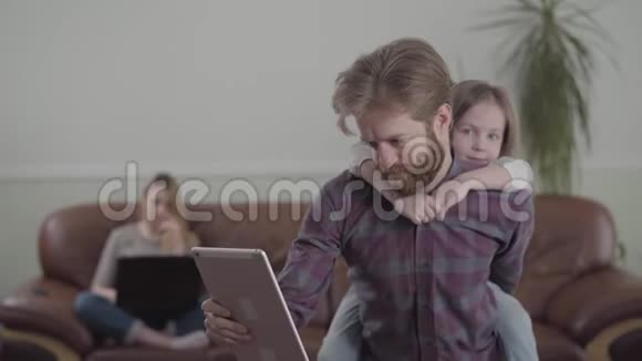 这位留胡子的人在平板电脑上工作参与了他的工作悲伤的小女孩坐在父亲的背上试图得到视频的预览图