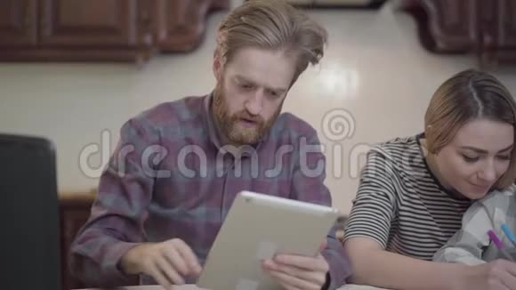 大胡子坐在桌旁手里拿着平板电脑漂亮的女人坐在旁边小女孩帮助女儿视频的预览图