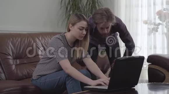 可爱的年轻女人坐在皮革沙发上胡须男站在她身边拿着笔记本电脑工作小女孩来了视频的预览图